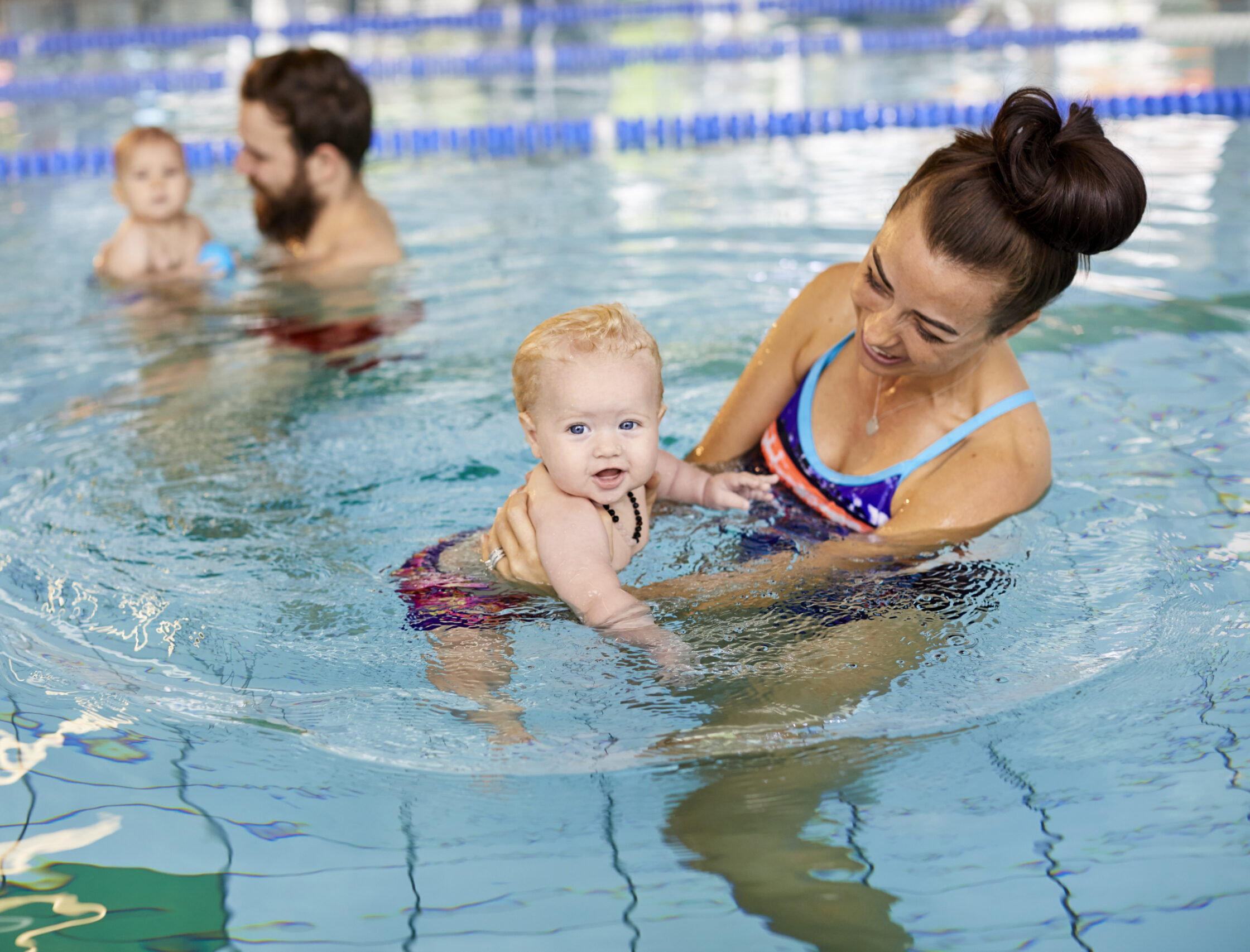 Photo of mother in the swimming pool, smiling whilst holding and looking at her baby in the water. A father and baby are nearby in the background.