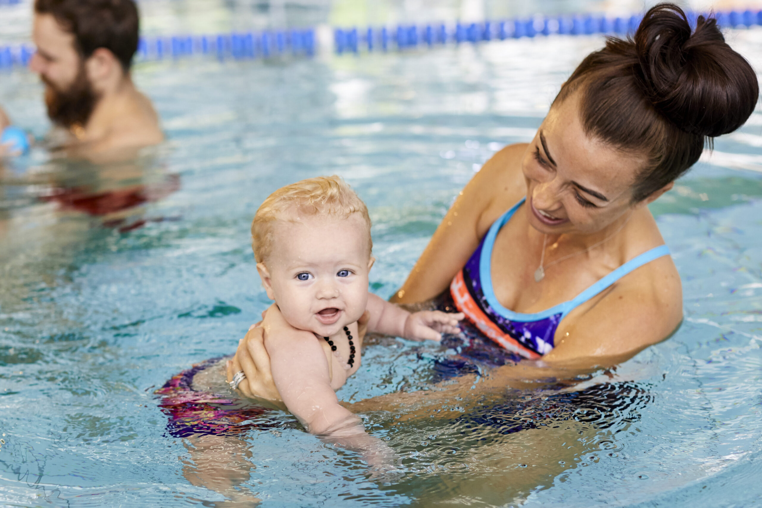 Photo of mother in the swimming pool, smiling whilst holding and looking at her baby in the water. A father and baby are nearby in the background.